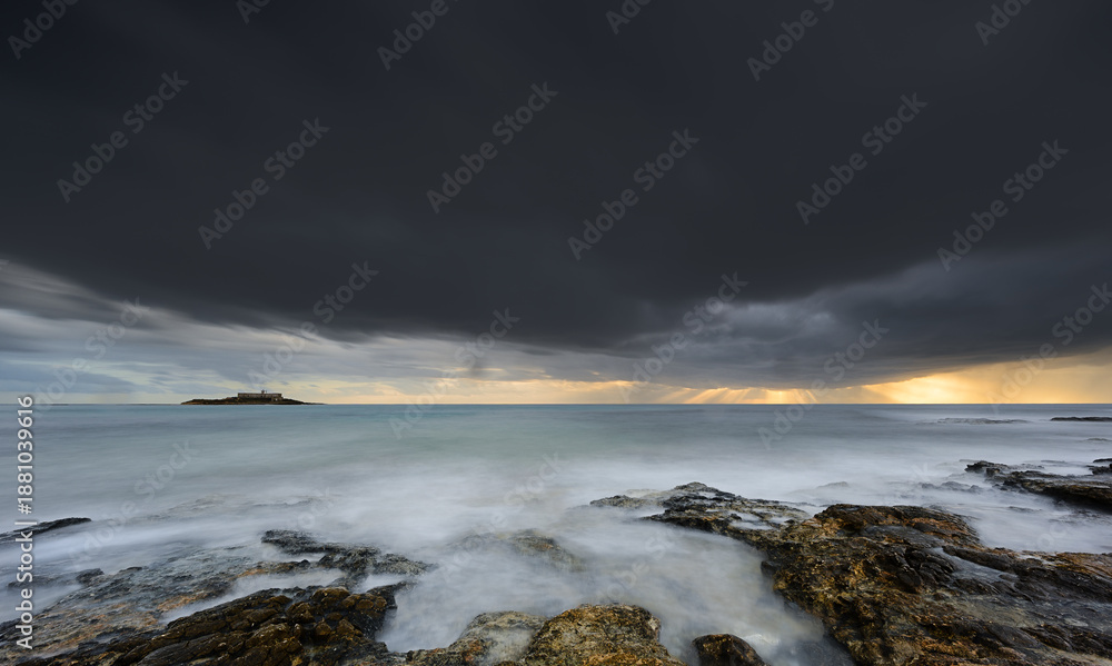 Obraz premium A stormy ocean with a rocky shoreline. The sky is dark and cloudy, and the water is choppy