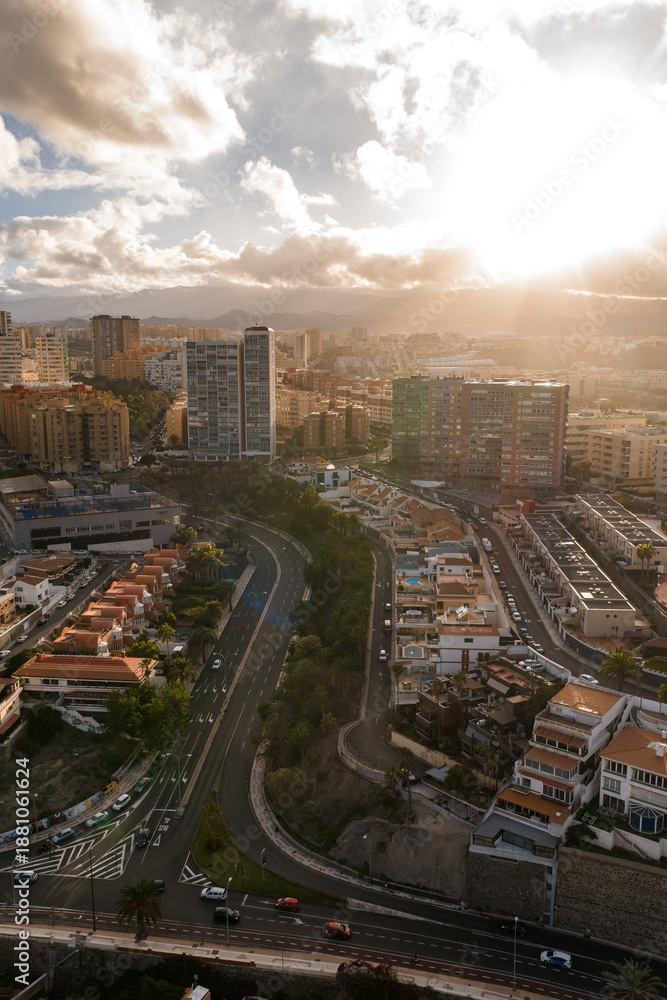 Fototapeta premium Aerial cityscape in Las Palmas de Gran Canaria shows S curve multilane roads, high rises, terraced blocks, winter sun rays, mountains, and traffic with pedestrians moving.