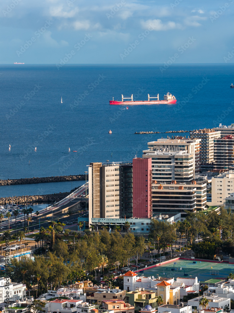 Fototapeta premium Las Palmas de Gran Canaria port scene shows mid and high rises, a red cargo ship offshore, curving breakwaters, sailboats, tennis courts, and palm lined streets in soft afternoon light.