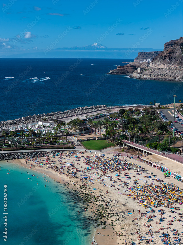 Fototapeta premium Aerial scene shows Amadores Beach, Gran Canaria, with umbrellas, promenade, palm park, marina, rocky headlands, and distant Mount Teide under bright midday light.