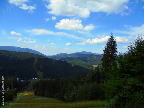 Wide view across rolling hills of the Beskydy Mountains with dense conifer forest, a cleared ski slope, and scattered meadows. Daytime light with fair-weather cumulus clouds and deep blue sky.