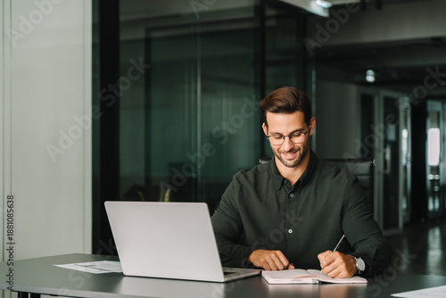 Smiling young man student using laptop computer for business studying watching online webinar e-learning training meeting writing notes. European businessman entrepreneur working in office, copy space