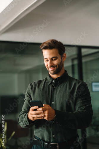 Smiling latin Hispanic business manager using cell phone mobile app. European young student man businessman holding smartphone standing in office or university, working online on device. Vertical