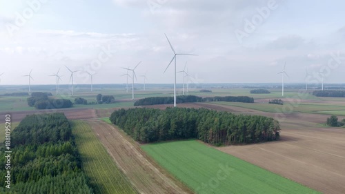 Aerial view shows white onshore wind turbines over patchwork fields and pine stands. Blades rotate slowly as a drone sweep reveals spacing and a narrow service track.