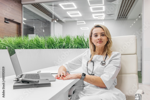 Εκτύπωση καμβά Female doctor in white medical attire is seated at a modern desk with a laptop,