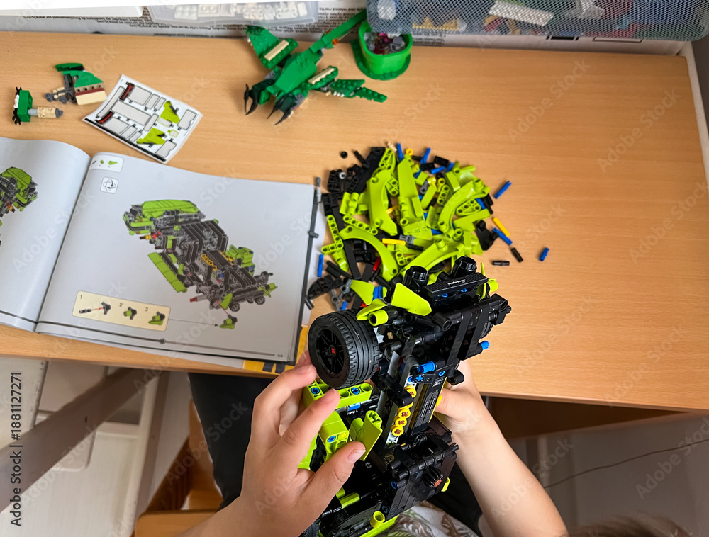 Naklejka premium A child's hands holding a partially assembled LEGO Technic car model, following the instruction manual on a des, flatlay: 04.05.2024- Ljubljana, Slovenia.