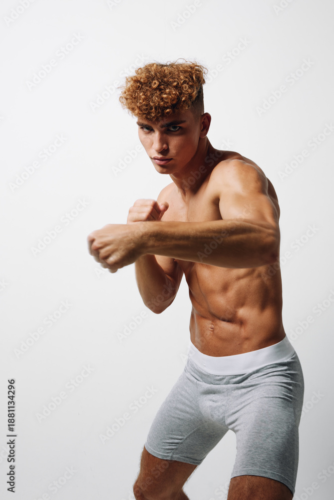 Fototapeta premium Athletic young man with curly hair practicing boxing punch in gray shorts against white background. Muscular fit male training for fitness and combat sport workout.
