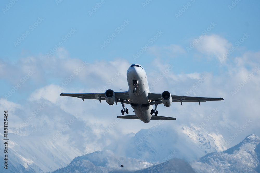 Naklejka premium Takeoff with Stunning Mountain Backdrop. White airplane taking off against snow-capped mountains, clouds and a blue sky.
