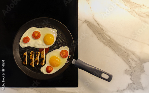 Fotografija Frying pan with sunny side up eggs and cherry tomatoes on a modern induction cooktop
