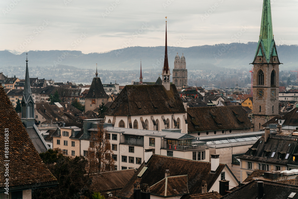 Fototapeta premium A panoramic cityscape of Zurich featuring historic church towers and rooftops under a cloudy sky. The image captures the charm of Switzerland old town with mountains in the background.