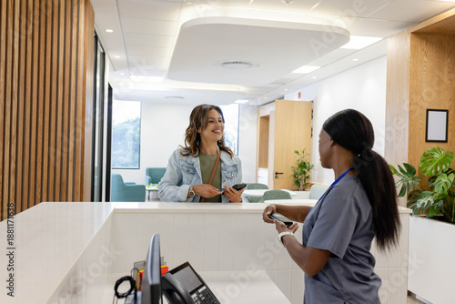 African American women at desk talking, denim jacket visitor, scrubs staff holding phone and card