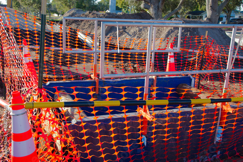 Construction Site With Trench Wall Supports, Orange Safety Netting, Barriers, and Piles of Dirt in Work Zone. Enclosed by orange safety netting and metal barriers, with dirt mounds and construction el