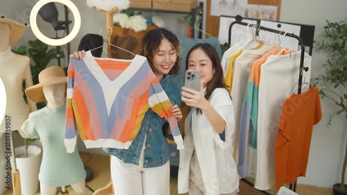 Two enthusiastic Asian women entrepreneurs create engaging social media content for their online clothing business, taking a cheerful selfie video while showcasing colorful striped sweaters to client