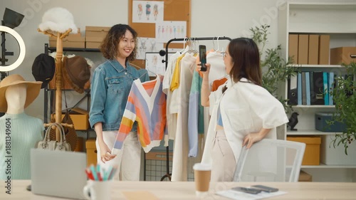 Two enthusiastic Asian women entrepreneurs create engaging social media content for their online clothing business, taking a cheerful selfie video while showcasing colorful striped sweaters to client