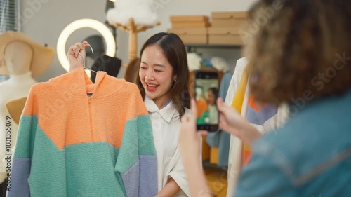 Two enthusiastic Asian women entrepreneurs create engaging social media content for their online clothing business, taking a cheerful selfie video while showcasing colorful striped sweaters to client