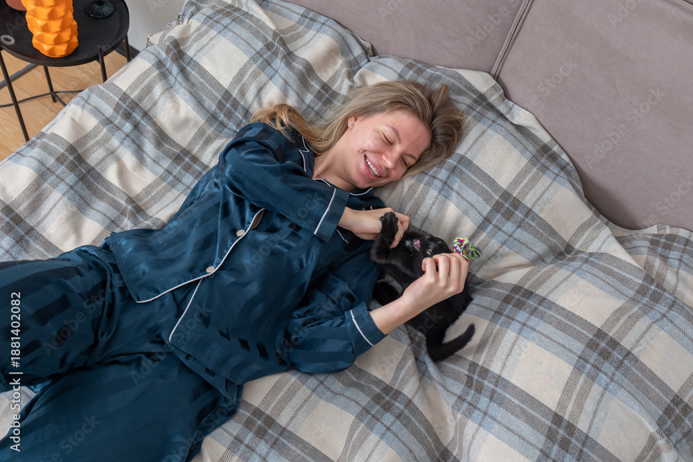 Fototapeta premium Smiling woman entertaining playful kitten indoors with cat wand toy, comfortable home lifestyle scene showing pet care bonding and leisure time.