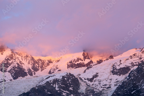 Warm pink and orange alpenglow lights up the snow-covered peaks and rugged ridges of Mont Blanc du Tacul, with soft clouds drifting across the dramatic alpine landscape.