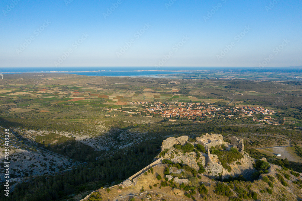 Obraz premium Ancient stone remains of Chateau de Opoul Perillos overlook a mosaic of farmland and a distant village, with the blue sea shimmering on the horizon under clear skies.