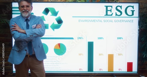 Confident senior businessman standing with arms crossed in front of large ESG presentation screen showing charts and sustainability data during corporate meeting in modern eco conference office.