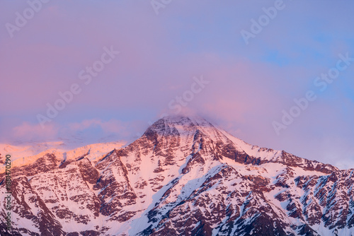 The snow-covered peak of Mont Blanc is partially obscured by soft pink clouds at dusk, with gentle evening light casting a pastel glow over the rugged alpine slopes.