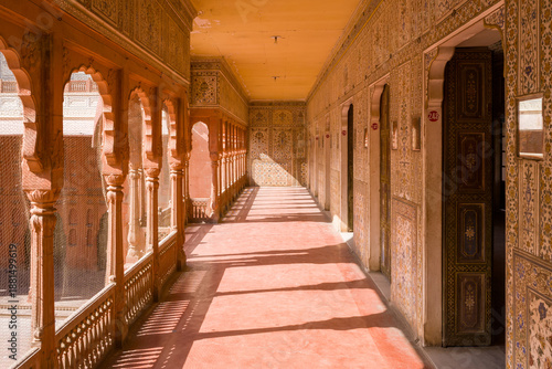Long corridor lined with ornate columns and decorative arches at Junagarh Fort, Bikaner, Rajasthan. Warm sunlight streams through the arches, casting intricate shadows on the red floor and