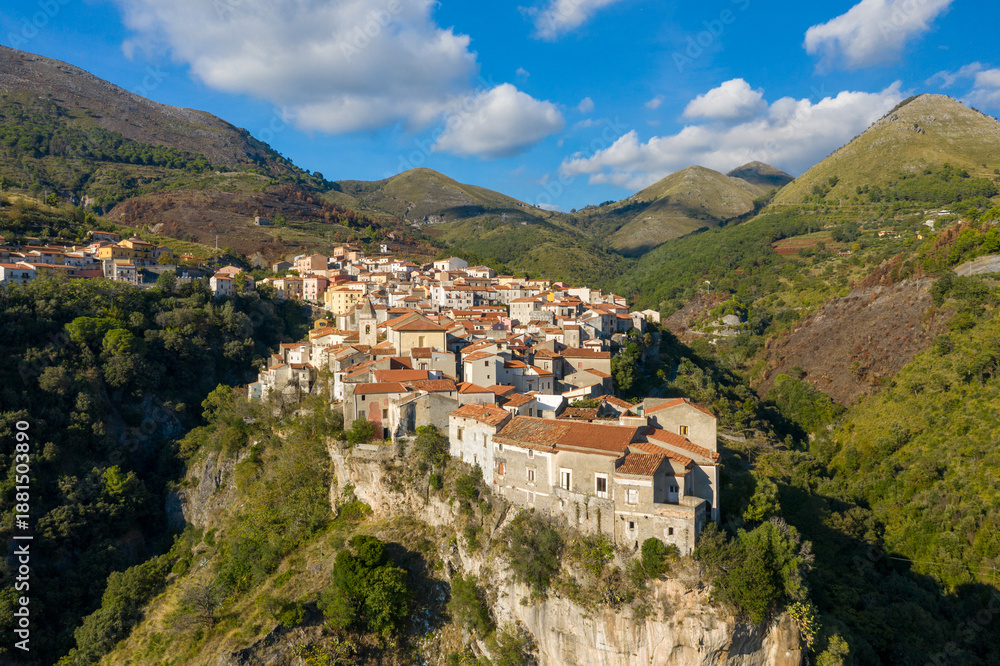 Obraz premium Vivid aerial view of Tortora, Italy, where rustic stone homes with red-tiled roofs cluster dramatically along the edge of a steep cliff, surrounded by rolling green hills under a bright blue sky with