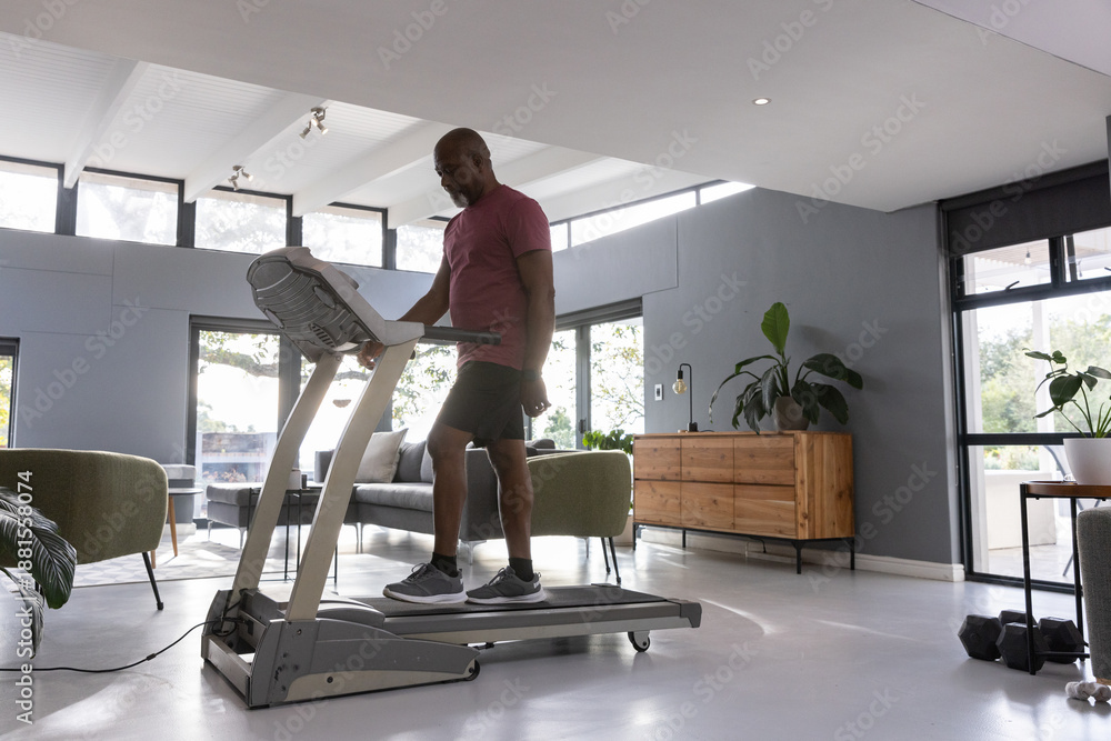 Fototapeta premium Man running on treadmill in living room flooded by natural light with sofa, dumbbells, plants