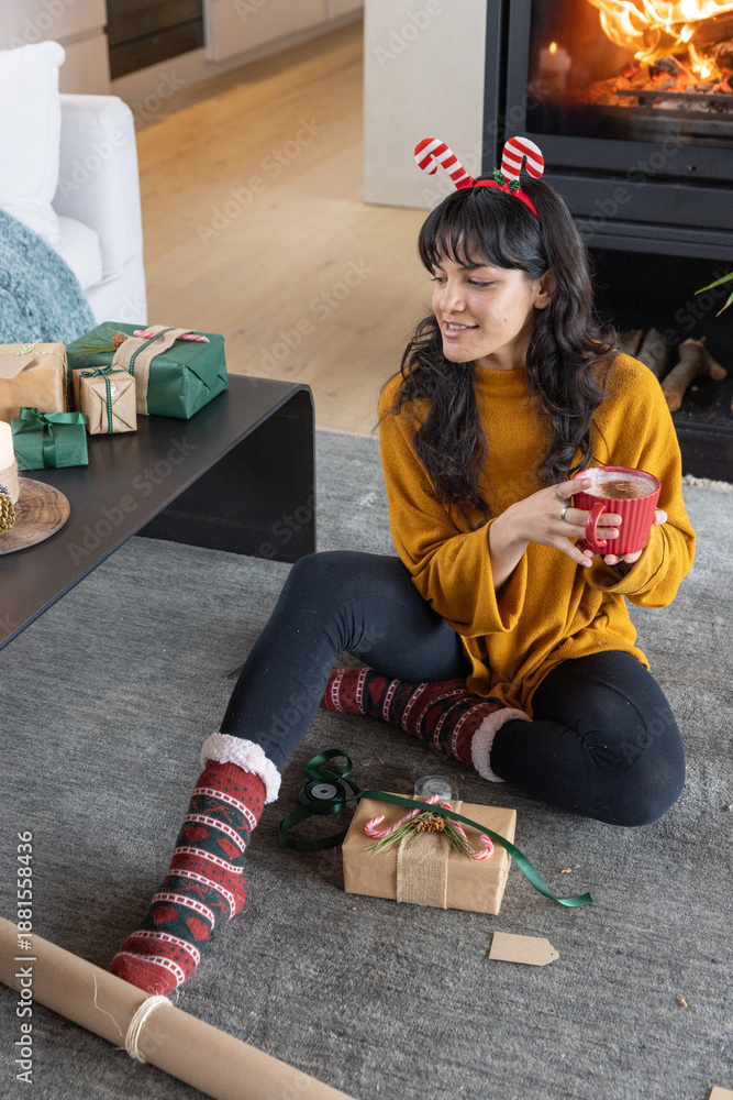 Fototapeta premium Asian woman in her twenties wearing mustard sweater in living room wrapping gifts, holding mug