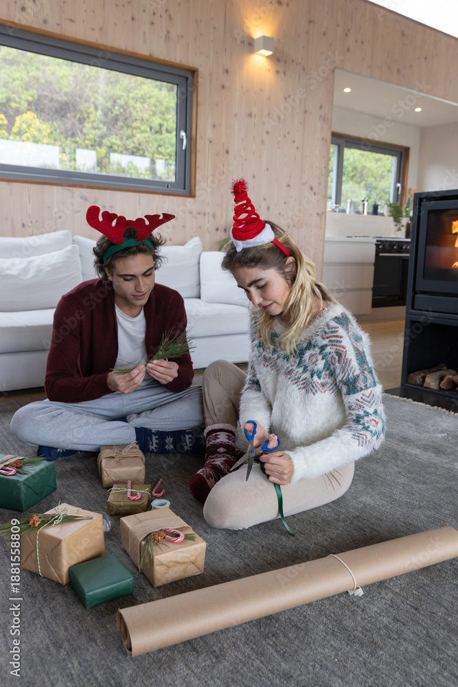 Fototapeta premium Couple sitting on gray rug in living room wrapping gifts with scissors, ribbon and kraft paper