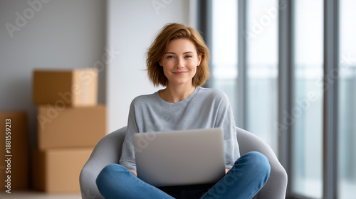Young woman smiling, sitting with laptop on her lap, using computer and connecting online while relocating or moving to new home, copy space