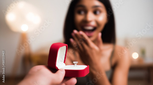 Hand presenting an engagement ring in a red box to a surprised woman with dark skin at home, symbolizing a marriage proposal, love, and future wedding plans