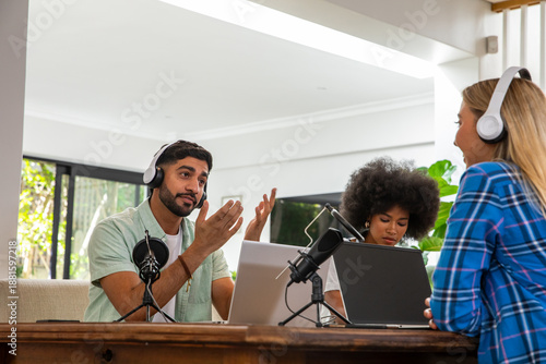 Diverse podcast hosts recording episode in home workspace with laptops and mic