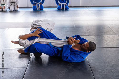 Blue and white brazilian jiu-jitsu gis grappling on padded mat in dojo, with kneeling gis