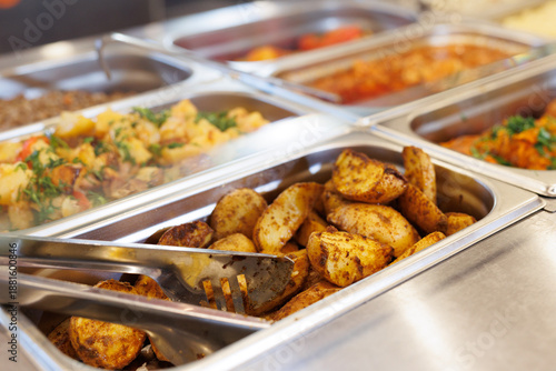 Potatoes in a buffet serving tray with tongs next to other dishes.