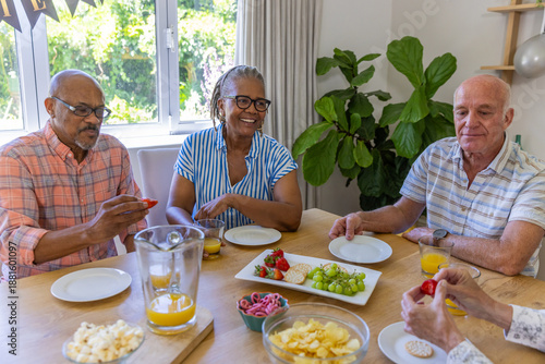 Diverse senior friends sitting around wood dining table at home, sipping orange juice, snacking