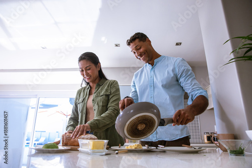 Diverse couple slicing bread and pouring eggs from frying pan at kitchen island with avocado