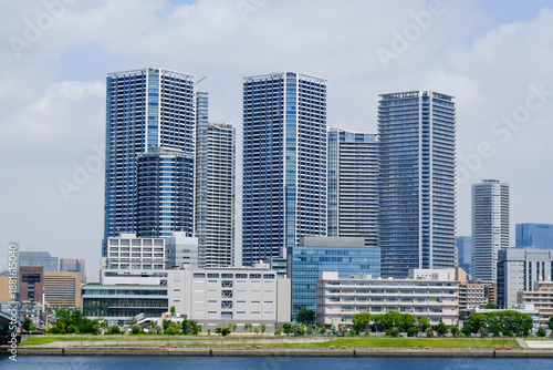 A wide-angle landscape photograph shows the skyline of a modern city along a river on a sunny day. Several tall skyscrapers with glass and concrete walls stand out in the right and center of Tokyo.
