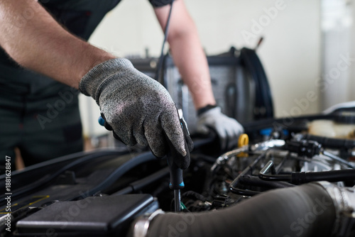 Man wearing work gloves repairing car engine in workshop, using screwdriver and focusing on mechanical components under open hood, demonstrating automotive maintenance skills