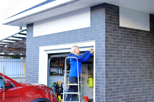 A senior Caucasian man with gray hair works on a garage door while standing on a ladder. A red car is parked nearby. The garage is organized with tools visible inside.