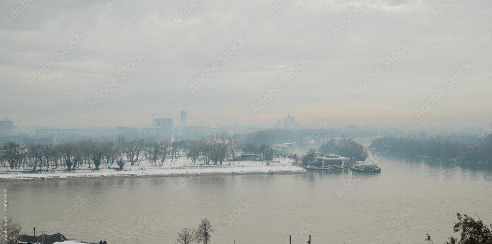 Fototapeta premium Winter river panorama of Belgrade with snowy riverbank, bare trees and misty city skyline, creating calm atmospheric urban landscape in cold season.