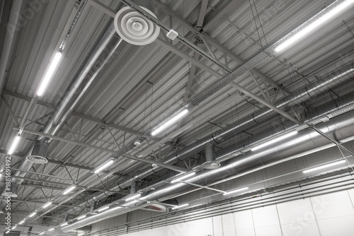 Wide view of an industrial ceiling featuring exposed HVAC ducts, ventilation vents, and suspended LED lighting in a large, clean workspace. Includes structural beams and copy space