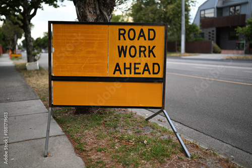 Yellow and black road works ahead sign, turned to face towards the footpath, on a suburban street late in the day