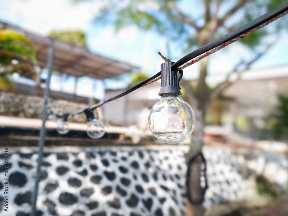 Obraz premium Close-up of outdoor string lights hanging in a backyard patio with a blurry garden background on a sunny day. Ideal for cafe, event, or home decor lifestyle concepts