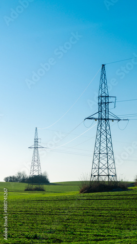 Two high-voltage power lines stand one behind the other in a green field against the backdrop of a blue sky on a sunny day.