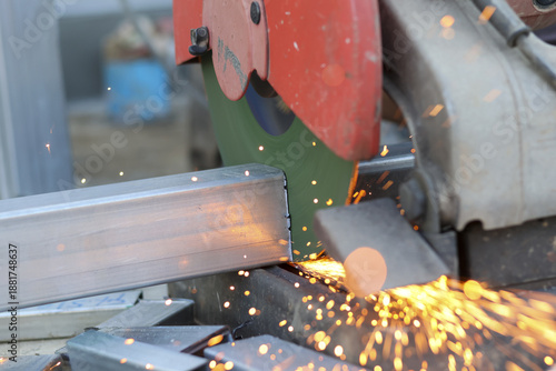 Energetic industrial workshop scene showing electric saw blade cutting steel bar with bright hot spark flying around creating dangerous intense atmosphere of heavy manufacturing process