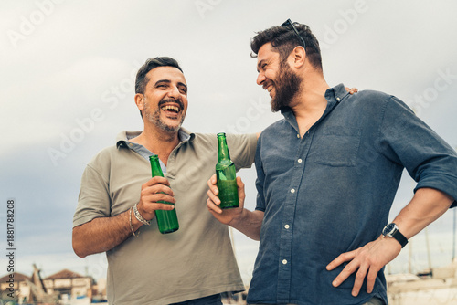 Two adult men laughing and drinking beer together outdoors