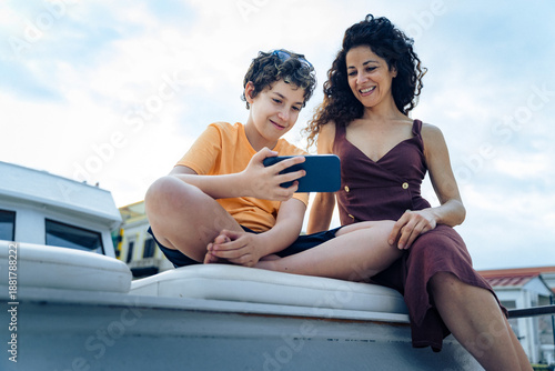 Mother and son using smartphone together outdoors on a boat - concept of family connection and tech