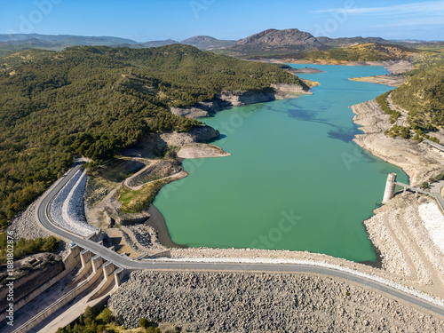 Aerial view of the turquoise waters of Embalse del Guadalhorce reflecting the clear sky, embraced by rugged mountains and the concrete curves of the dam, Ardales, Andalucia, Spain.