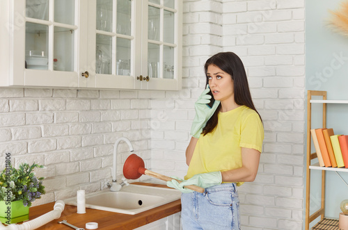 Young girl talking by the phone advising with plumber how to unclog a sink drain. Girl trying to clean sink using plunger standing in the kitchen at home. Housekeeping and household work concept.