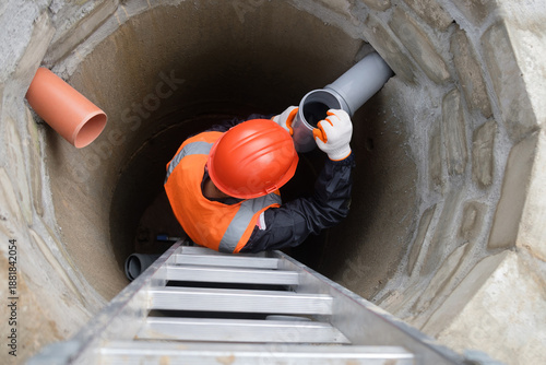 A worker wearing a safety helmet, protective gloves, and a high-visibility vest is installing or aligning a plastic sewer pipe inside a concrete sewage well of a private house. The image is captured f